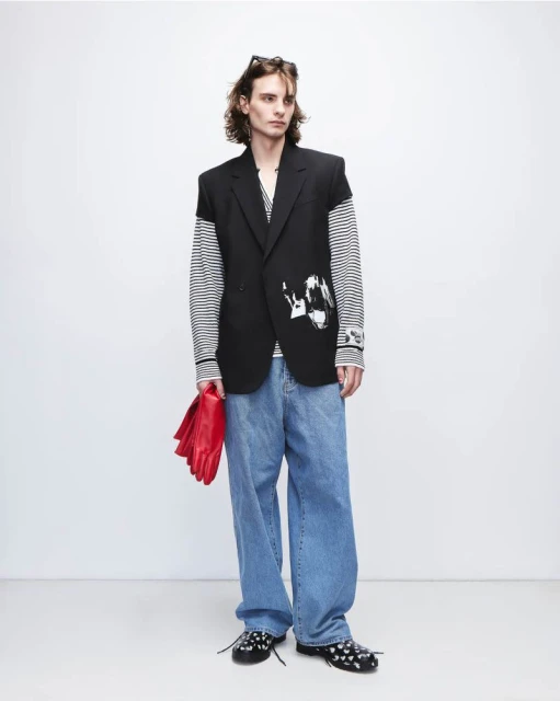 Full-length shot of a young adult male fashion model in an oversized black blazer, jeans, and black shoes against a white backdrop.