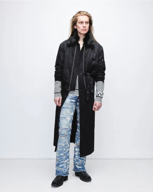 An adult male with long blonde hair is standing in a photo studio wearing a long black coat and ripped light blue jeans.