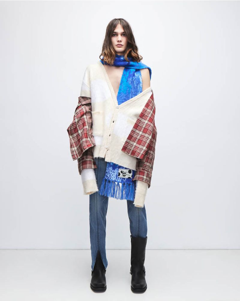 Full-length studio shot of a young adult male fashion model wearing a cardigan, plaid top, jeans, and boots in a bright, neutral space.