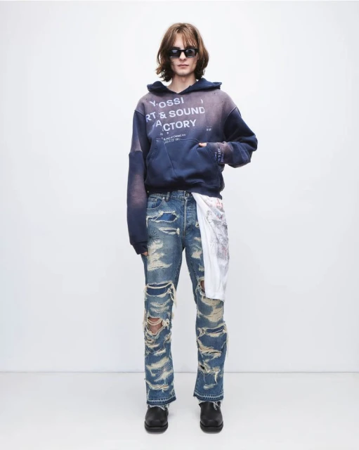 A long-haired man wearing sunglasses, a blue hoodie, and ripped jeans stands against a plain white backdrop in a fashion-style photo.