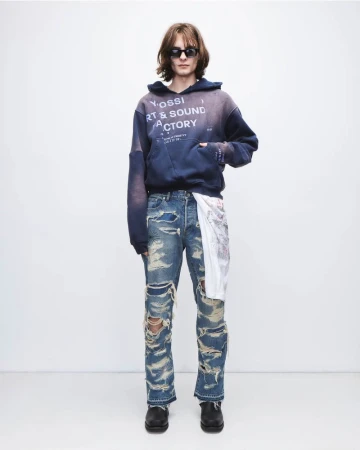 A long-haired man wearing sunglasses, a blue hoodie, and ripped jeans stands against a plain white backdrop in a fashion-style photo.