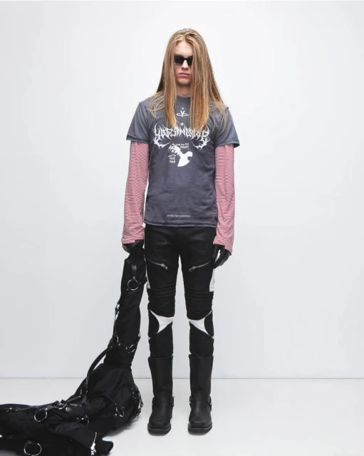 Full-length studio shot of a male model with long blonde hair, wearing a graphic tee, black pants, boots, sunglasses, and a black bag.
