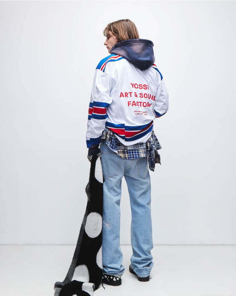 Adult male stands in white hockey jersey, jeans, and scarf. Back view in a photo studio setting. Fashion editorial photography.