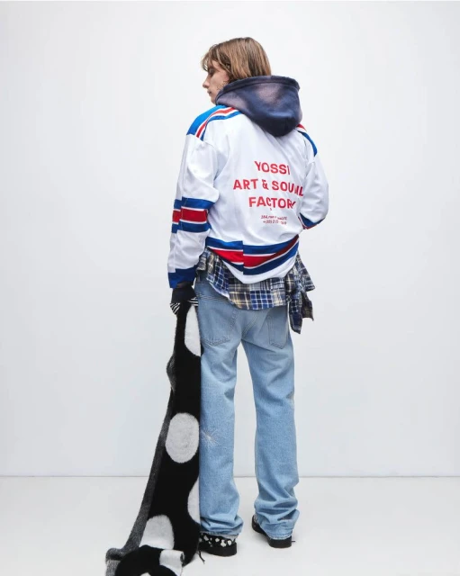 Adult male stands in white hockey jersey, jeans, and scarf. Back view in a photo studio setting. Fashion editorial photography.