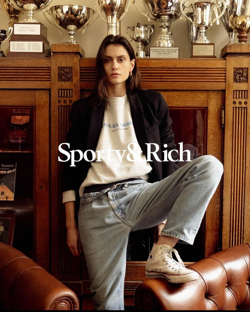 An adult female poses in front of a wooden cabinet in a grey sweatshirt, jeans, black jacket, and sneakers next to silver trophies.