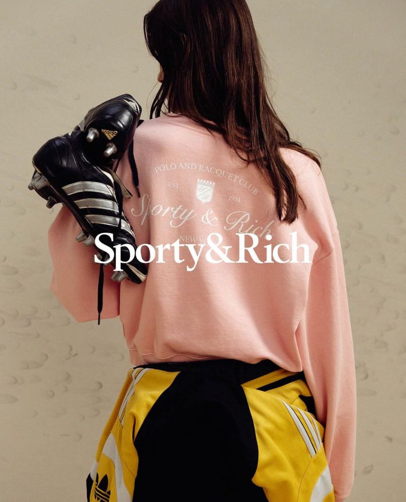 Back view of a woman in a pink sweatshirt and black and yellow athletic pants holding Adidas soccer cleats over her shoulder.
