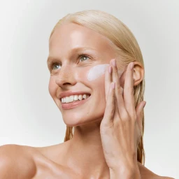 Close-up of a smiling blonde woman applying white skincare cream to her cheek against a neutral background.