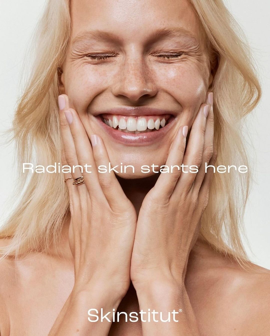 Close-up portrait of smiling blond woman with hands on face, advertising radiant skin, against a neutral background for skincare brand.