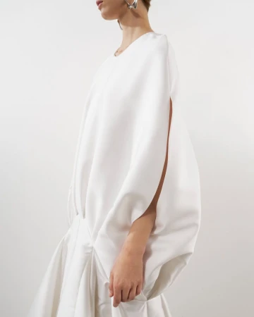 Model wearing a white sculptural dress with voluminous sleeves against a neutral seamless backdrop.