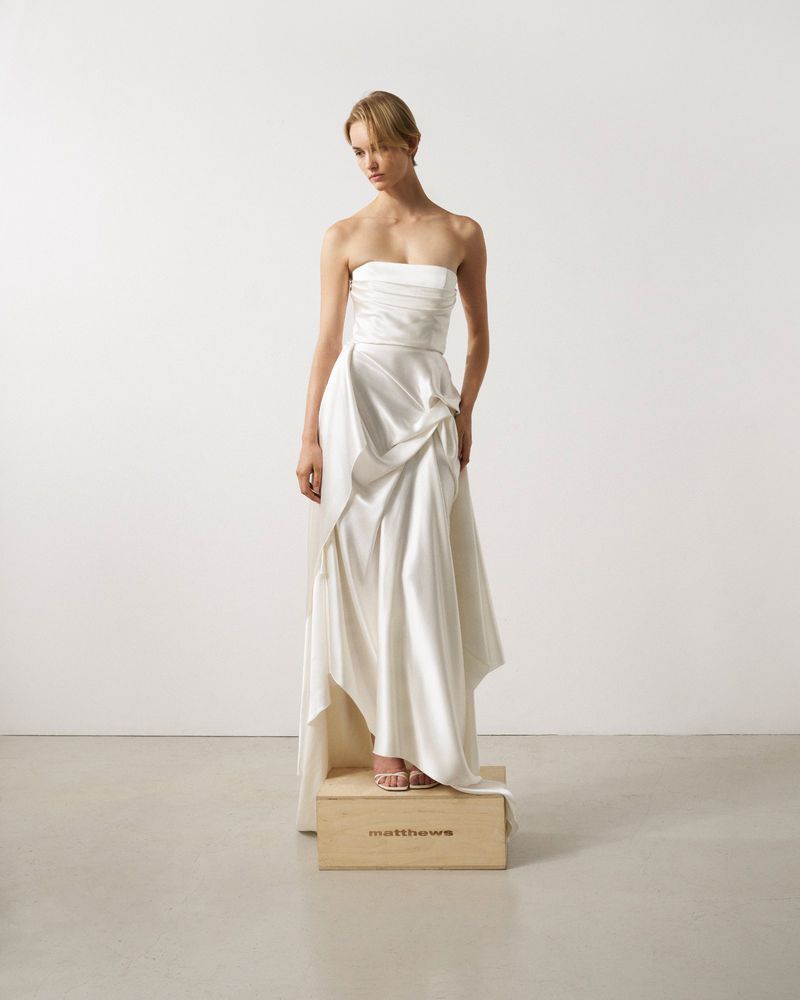 Woman in a strapless white gown standing on a wooden box in a minimalist studio setting. Soft, diffused lighting enhances the dress's texture.