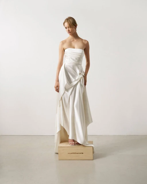 Woman in a strapless white gown standing on a wooden box in a minimalist studio setting. Soft, diffused lighting enhances the dress's texture.
