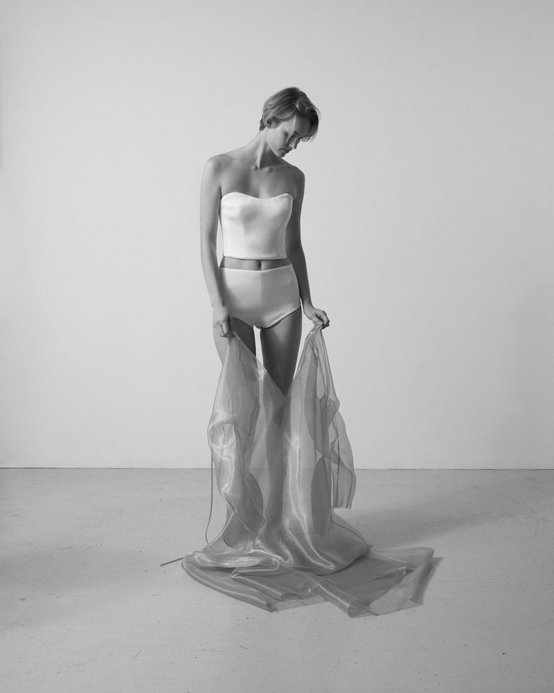 Black and white studio portrait of a slender woman wearing a corset and high-waisted briefs, holding a sheer flowing fabric.