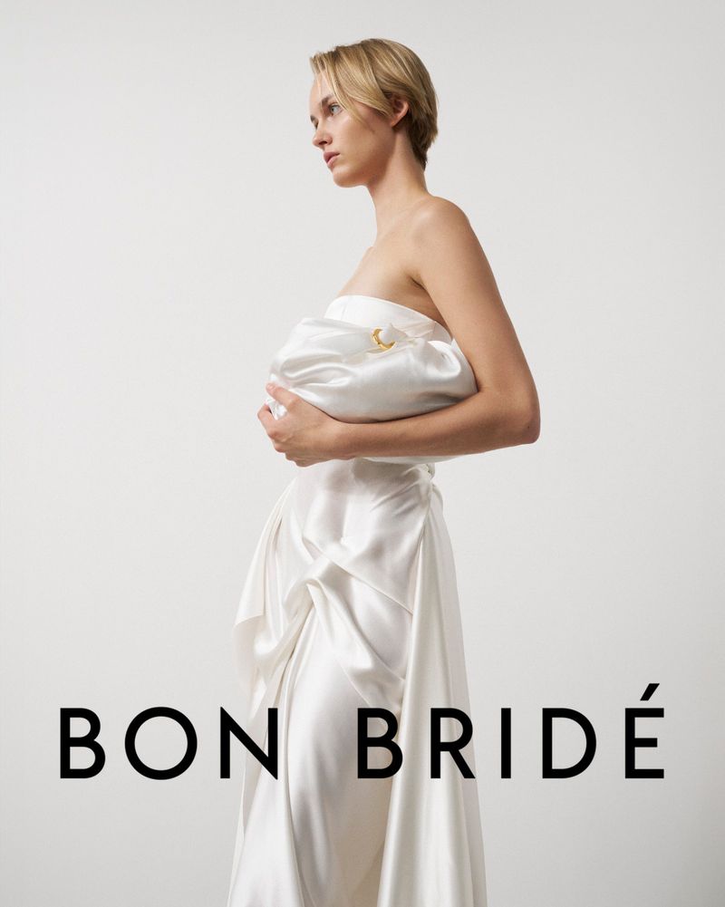 Elegant blonde woman in off-shoulder ivory dress holds matching clutch in a bright studio shot for a fashion editorial.