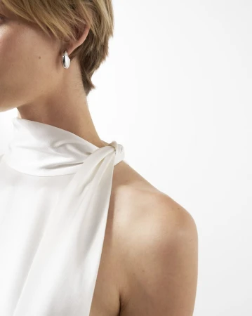 Elegant woman showcases a silver hoop earring against a white background, wearing a matching white top with a shoulder tie detail.