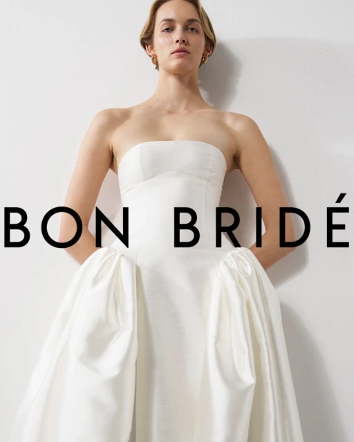 Elegant woman in a white strapless gown with a textured bodice and full skirt, posing in front of a plain white backdrop. Bridal fashion.