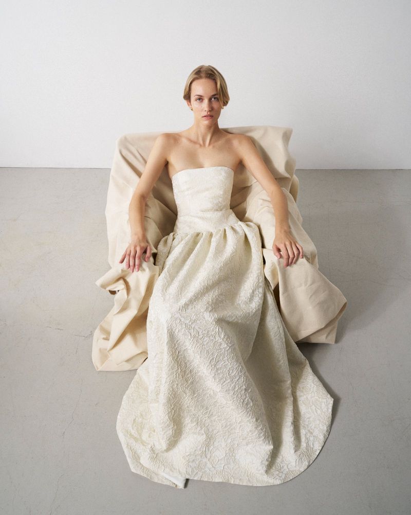 Woman poses in a strapless, textured gown while seated in a draped chair against a stark white studio backdrop, fashion editorial photography.