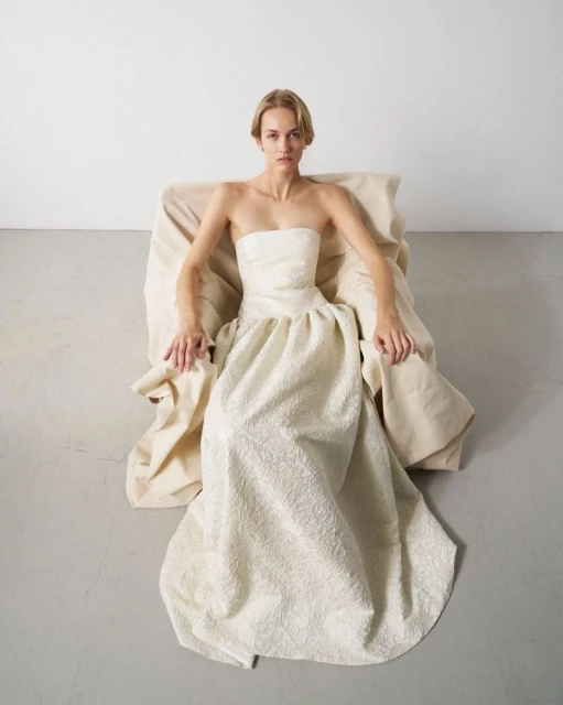 Woman poses in a strapless, textured gown while seated in a draped chair against a stark white studio backdrop, fashion editorial photography.