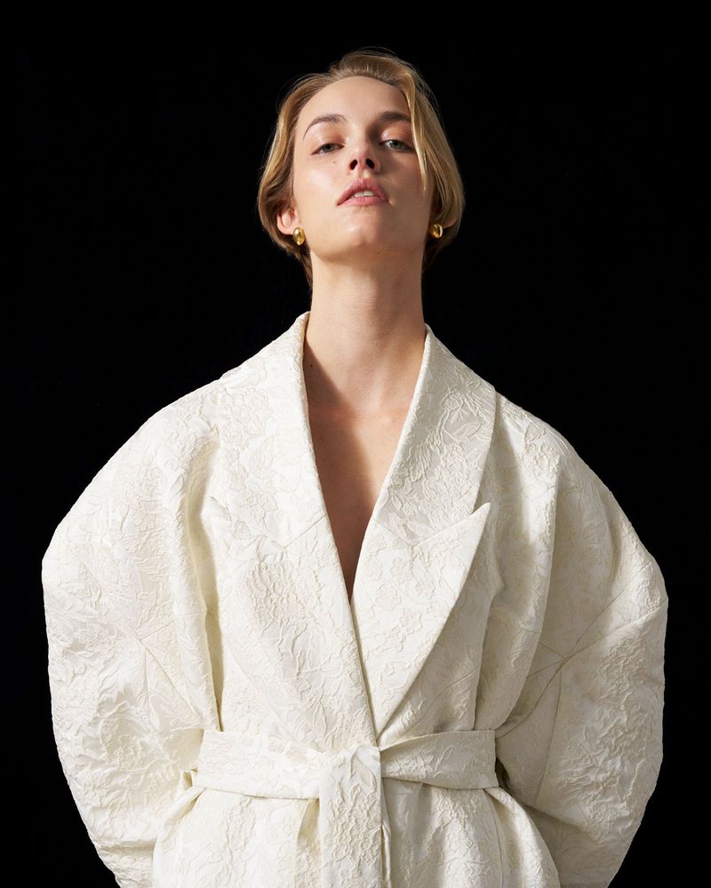 Blonde model in an off-white belted blazer posing against a black backdrop in a studio, looking directly at the camera.