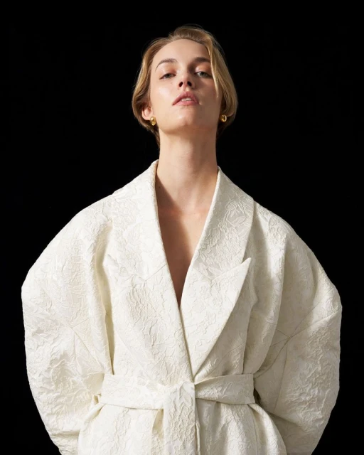 Blonde model in an off-white belted blazer posing against a black backdrop in a studio, looking directly at the camera.