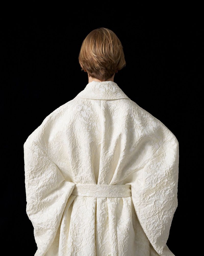 Back view of an adult female with short blonde hair wearing a textured, ivory-colored belted robe against a solid black background.