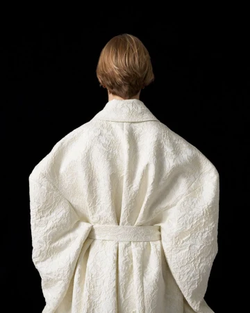 Back view of an adult female with short blonde hair wearing a textured, ivory-colored belted robe against a solid black background.