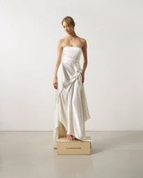 Model standing on a wooden crate wearing a strapless white silk wedding gown against a plain studio background.