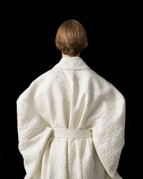 Model seen from behind wearing an oversized white textured belted coat against a black background.