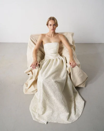 Model in a strapless white wedding dress seated in a draped armchair against a neutral studio background.