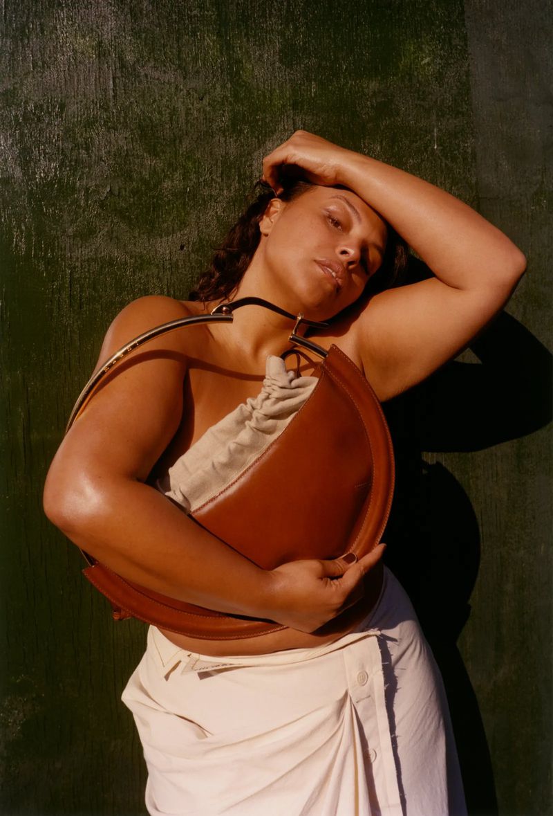 Woman with brown leather moon bag against a green wall. The woman has dark curly hair, wearing white draped skirt, holding the bag.