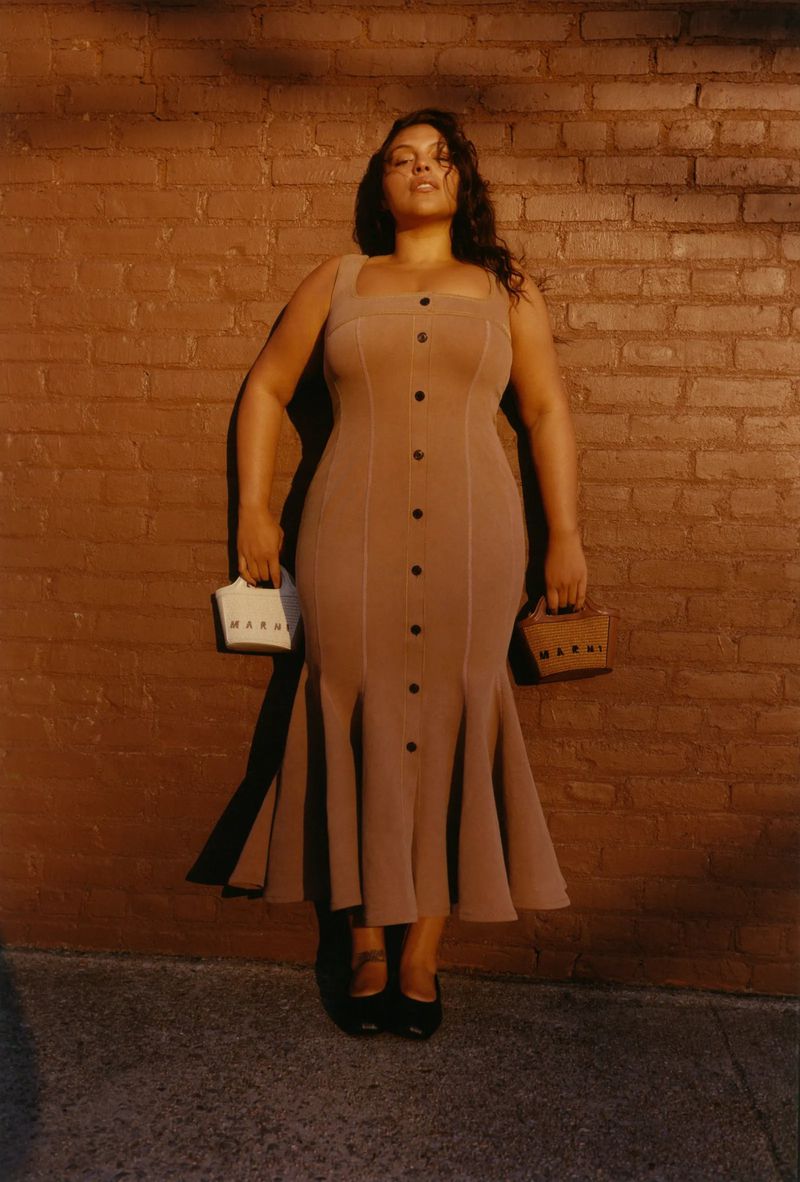Brunette woman in a taupe dress with button detail poses with two small box purses against a textured brick wall in outdoor lighting.
