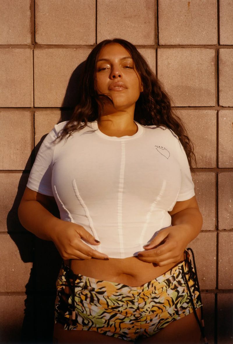 Woman with long dark hair poses in a white top and floral shorts against a textured tan brick wall in warm, natural light.