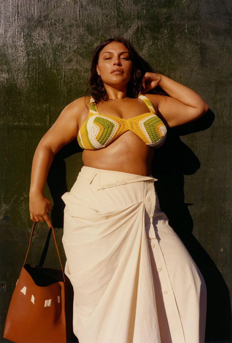 Woman in crochet bikini top and a cream wrap skirt poses with a brown tote bag against a green wall in a medium shot.
