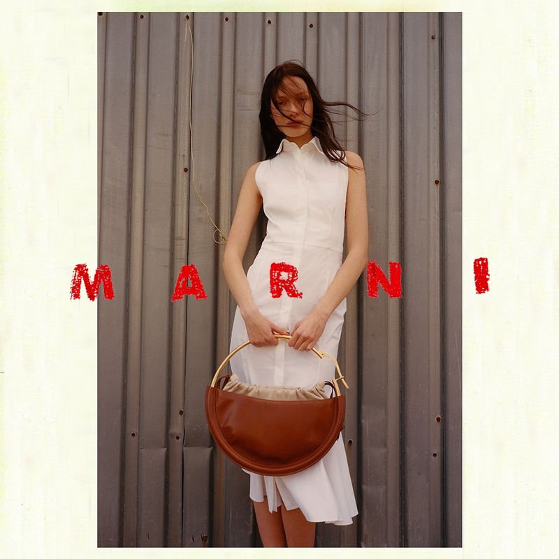 Fashionable woman in a white dress holds a tan leather handbag with a gold handle against a grey corrugated metal wall.