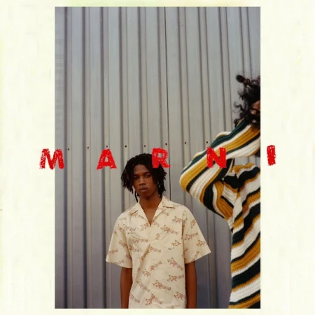 Two models pose indoors against a corrugated metal wall, one in a patterned shirt, the other in a striped sweater, for a fashion editorial.