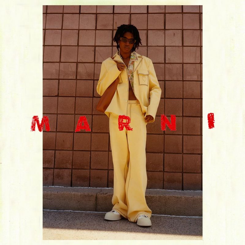 A person wearing a yellow suit stands in front of a brick wall with the word 'MARNI' superimposed in red letters.