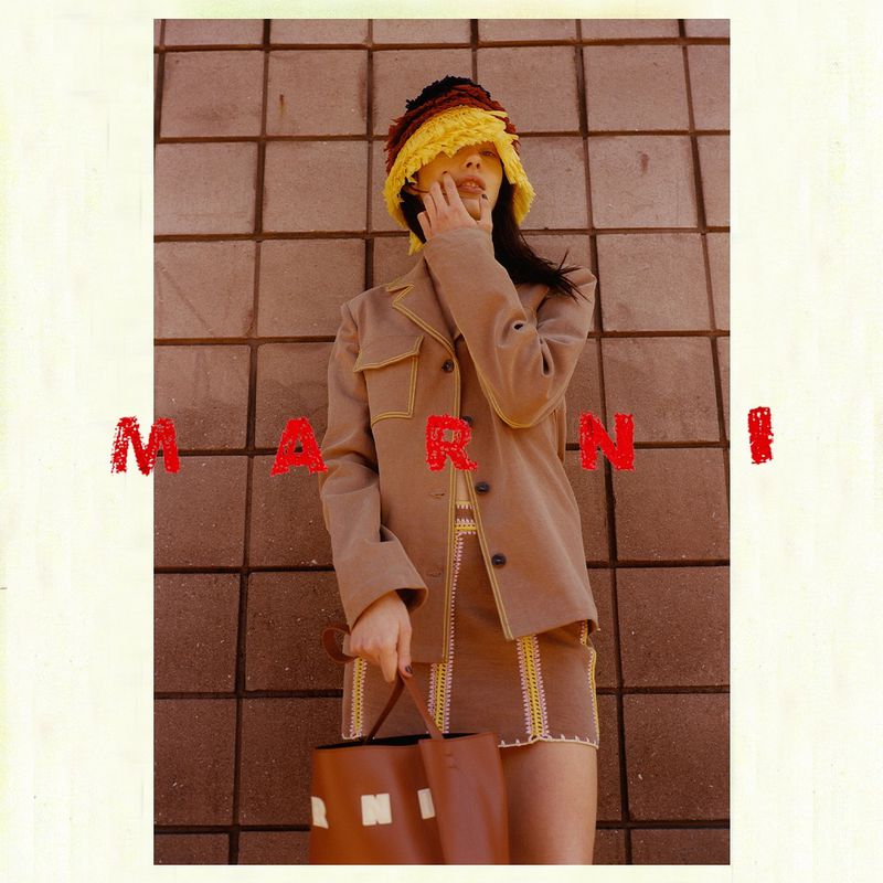 Indoor shot of an adult female model wearing a brown skirt and jacket with a colourful bucket hat, holding a leather bag and posing.
