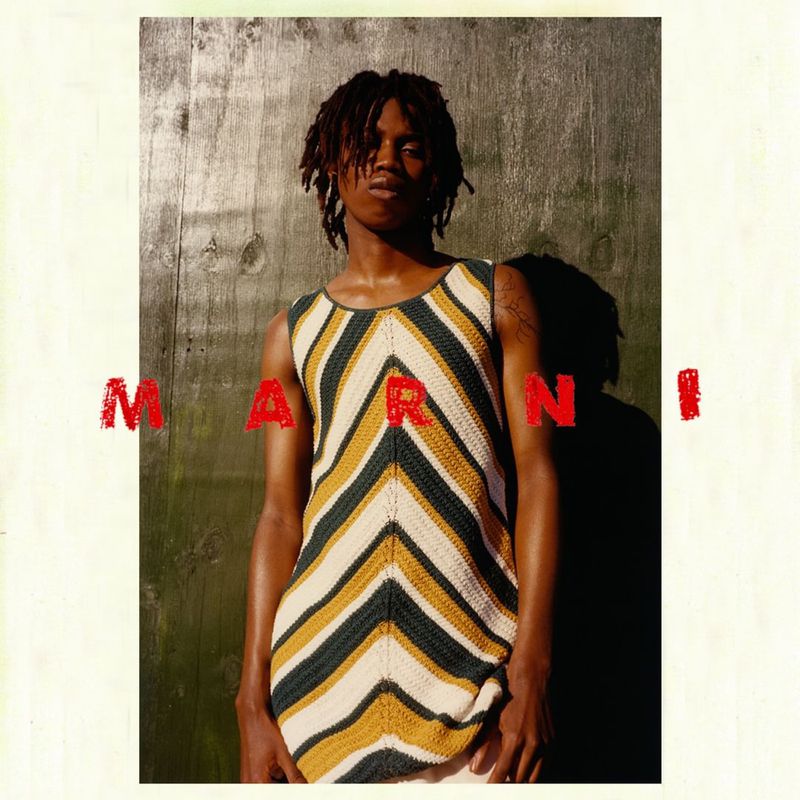 Male fashion model poses in a yellow, white, and green chevron patterned top, photographed against a weathered wooden wall indoors.
