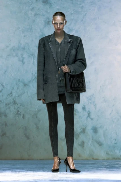 A woman posing in an oversized grey denim jacket, leggings, and heels, holding a black logo bag, standing in front of a textured background.