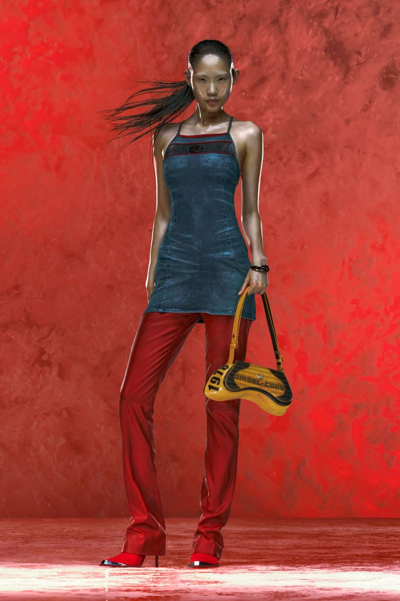 A female fashion model with black hair in a denim dress and red leather pants with a yellow purse poses against a red backdrop.