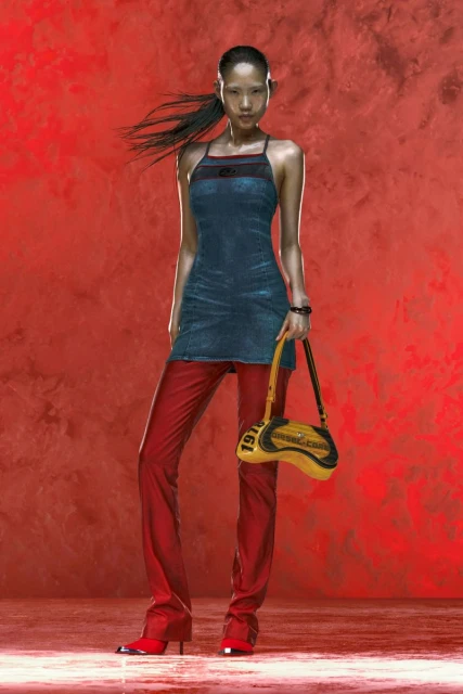 A female fashion model with black hair in a denim dress and red leather pants with a yellow purse poses against a red backdrop.