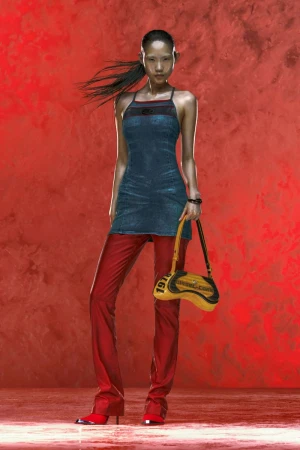 A female fashion model with black hair in a denim dress and red leather pants with a yellow purse poses against a red backdrop.