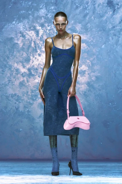 Fashion shoot featuring a full-length view of a model in a blue dress holding a pink purse against a textured, light blue background.