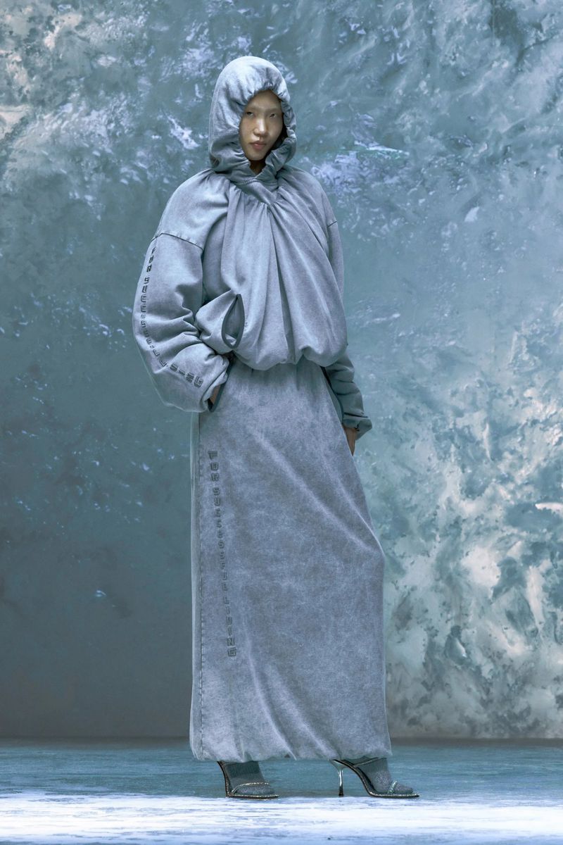 An Asian woman stands in a gray hooded outfit with long sleeves and a floor-length skirt, posing in front of a textured gray background.