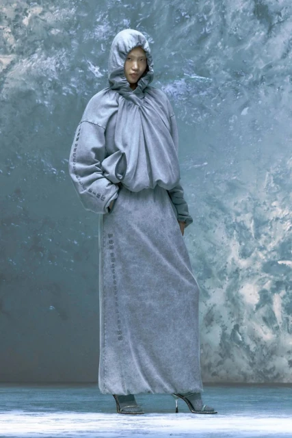 An Asian woman stands in a gray hooded outfit with long sleeves and a floor-length skirt, posing in front of a textured gray background.