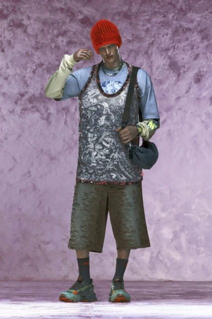 A person in a patterned tank top, shorts, and a red beanie stands in front of a textured purple and pink background, showcasing unique streetwear fashion.