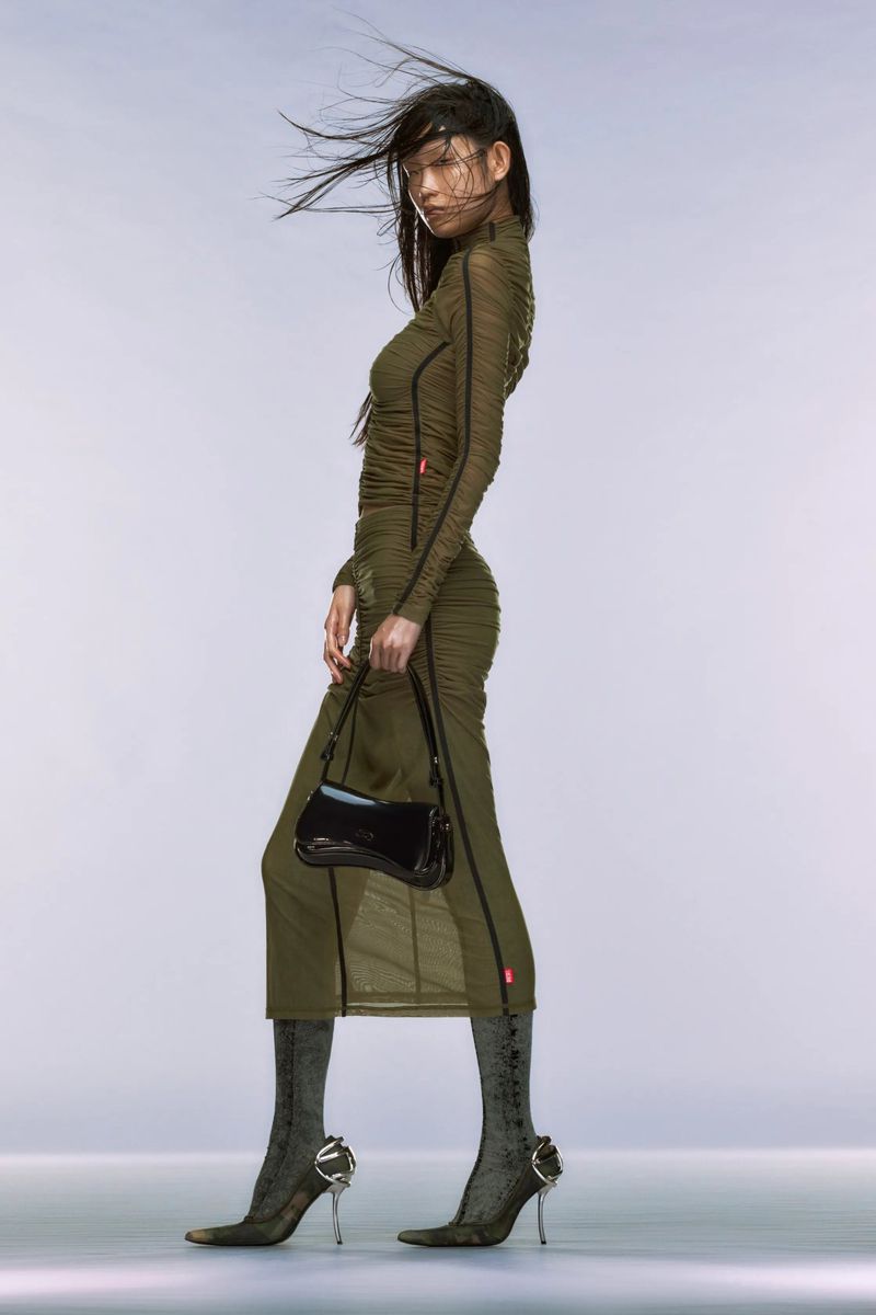 A fashionable Asian female model poses in an olive green dress, shiny black purse, and unique heels in a modern, high-fashion studio shot.