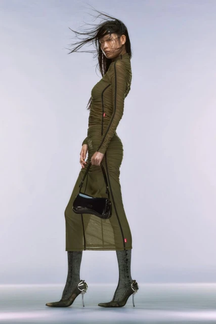 A fashionable Asian female model poses in an olive green dress, shiny black purse, and unique heels in a modern, high-fashion studio shot.