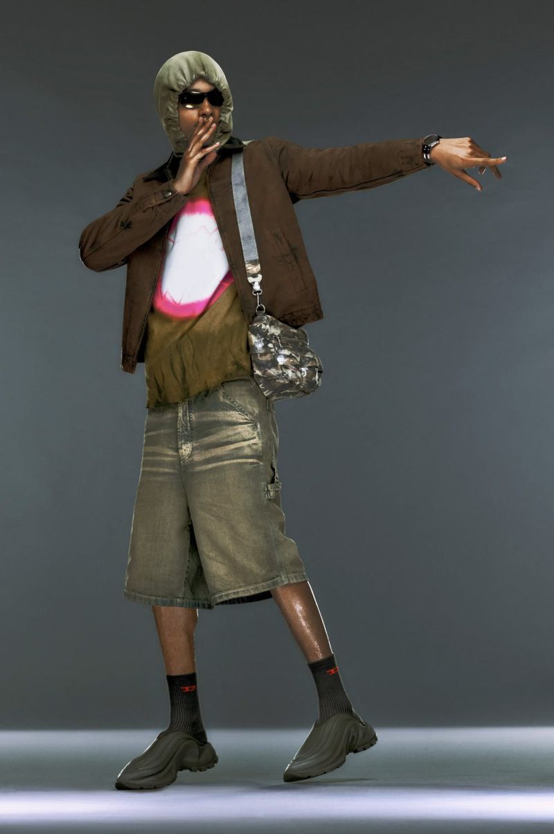 Full shot of a black male model in a brown jacket, faded denim shorts, sunglasses, and grey balaclava, posing in front of grey background.