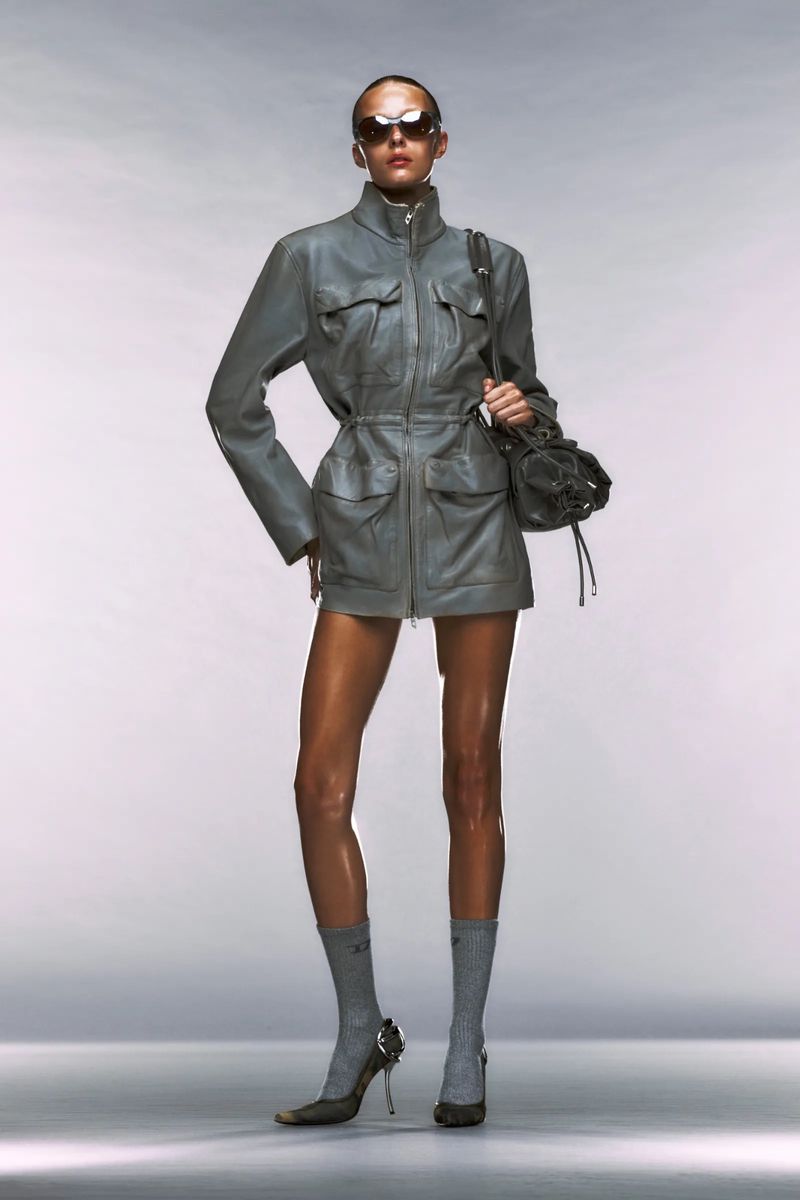 A fashion model poses in a grey jacket and heels, holding a soft black handbag in a studio shot with a clean white background.