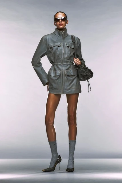 A fashion model poses in a grey jacket and heels, holding a soft black handbag in a studio shot with a clean white background.